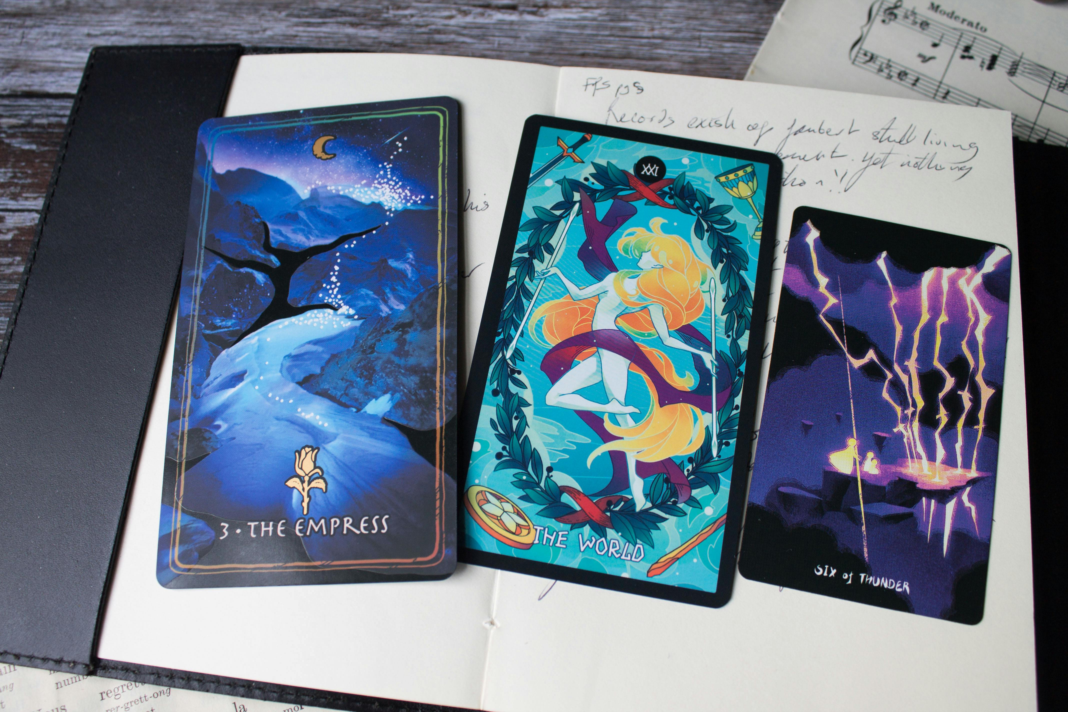 Three Tarot card from three different decks sit on top of an open Journal.  The first a dark night scene with a path of light leading from the flower at the bottom deeper and up to a beautiful starry sky. The Text below the flower is "The Empress". In the middle is an elemtental slyph dancing with two twirling sticks, one in each hand. Around it is a wreath and  a sword, cup, coing and wand, the Text at the bottom reads "The World". The final smaller card depicts 6 arcs of lightning striking an outcropping in front of 3 glimmer walkers. 2 are huddled together and the third stands behind them with a long pole of light. The text on this card reads "six of thunder". 