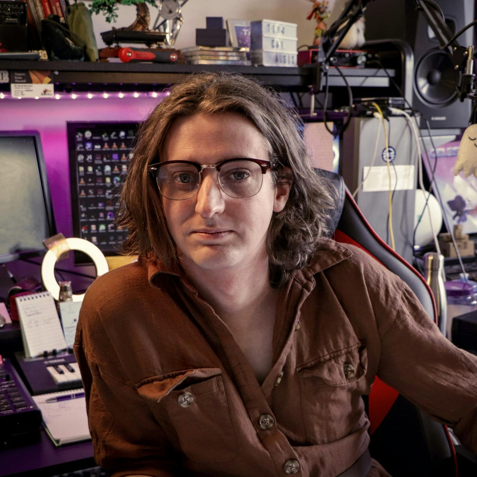  A photo of Amber, they're a white person with glasses and shoulder length curly brown hair. They're sitting in their studio with many gadgets and their desk behind them