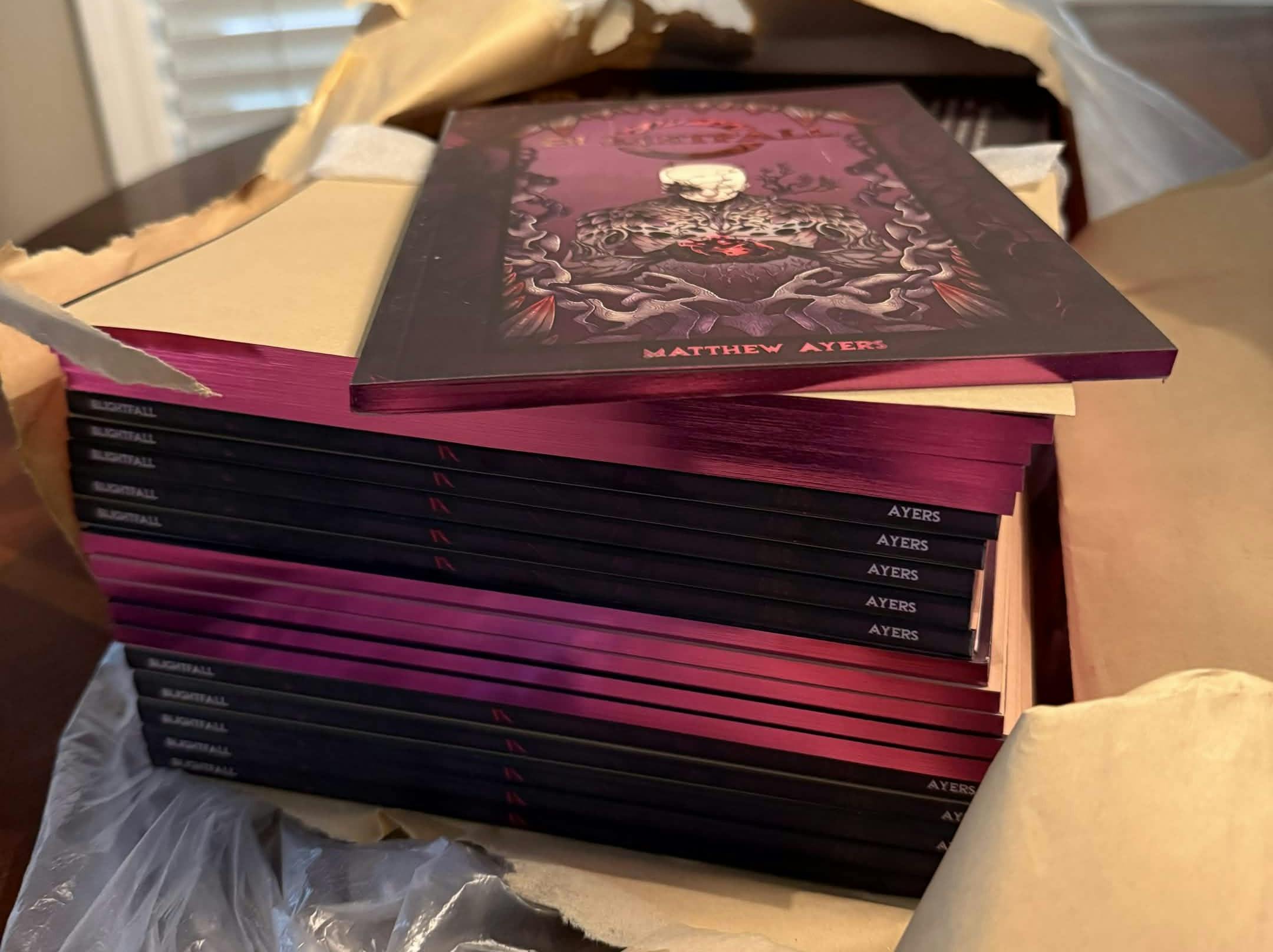 A pile of Blightfall physical books, copies alternating to show spine and painted edges in metallic purple
