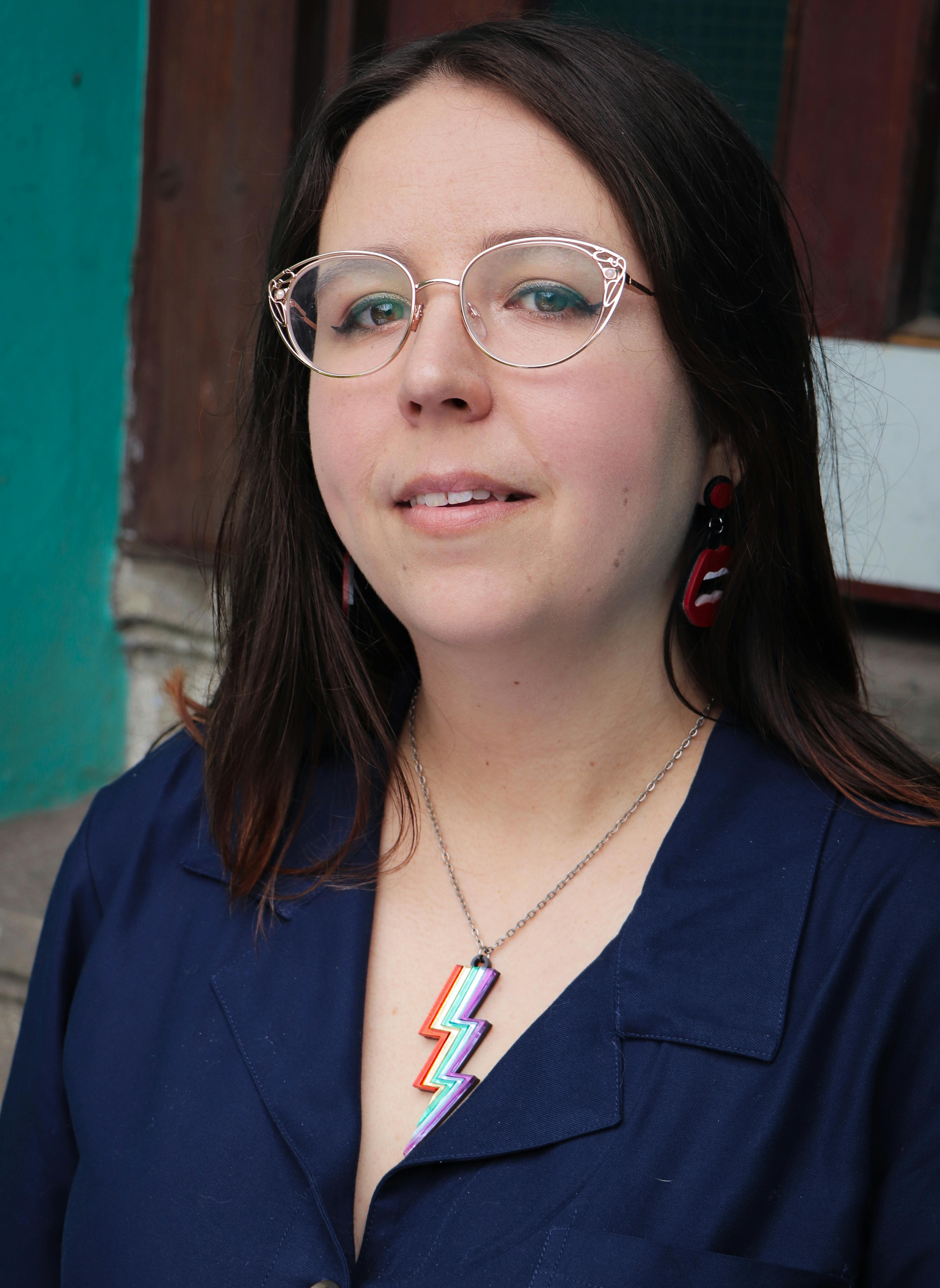  A photo of Ella, she's a white woman with brown hair and glasses, looking curiously at the camera. She's wearing a rainbow lightning bolt necklace 