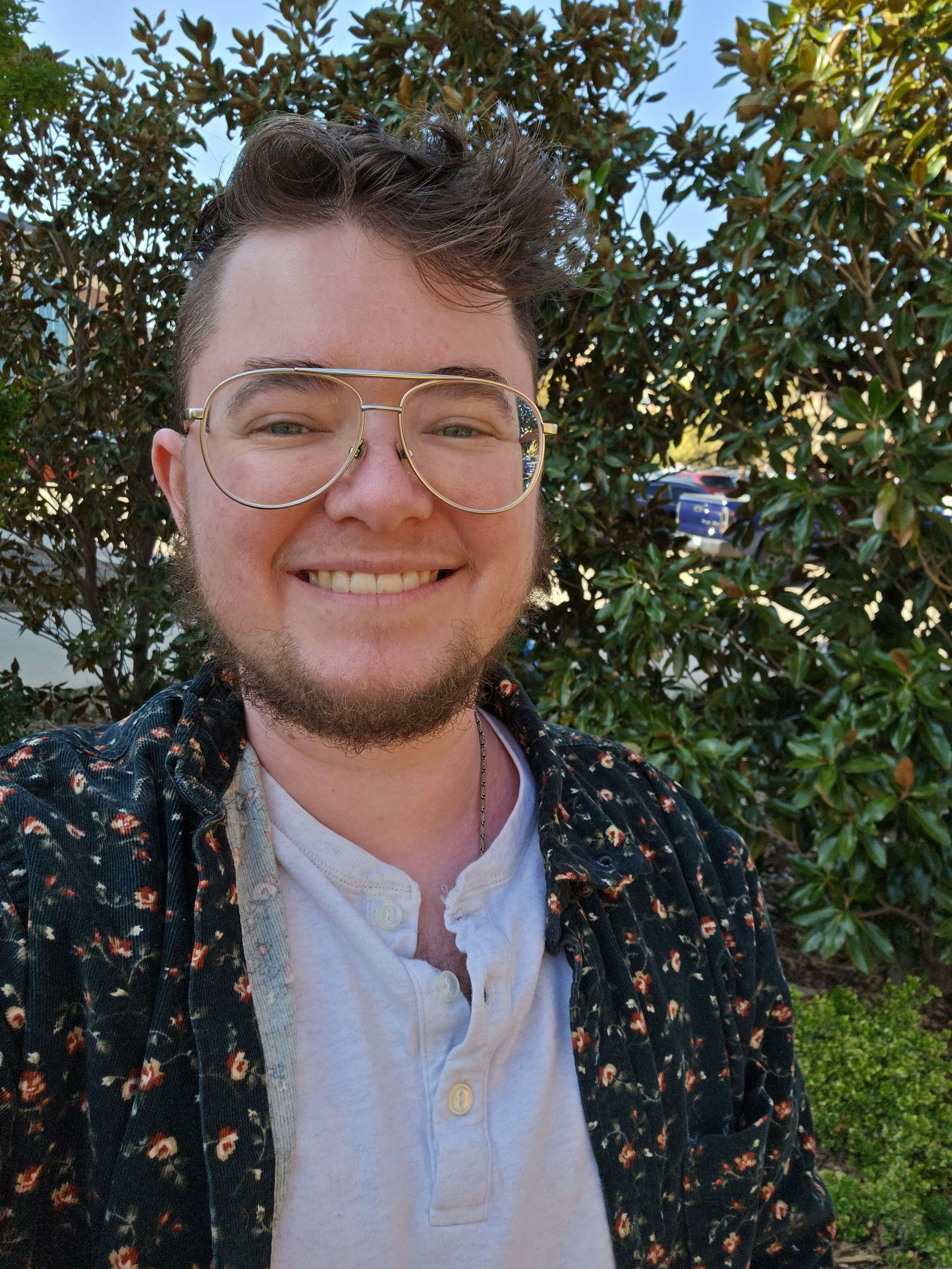  A photo of Luka, they're a pale person with glasses and a beard. He's standing outside smiling, wearing a flowery shirt over a white t-shirt.