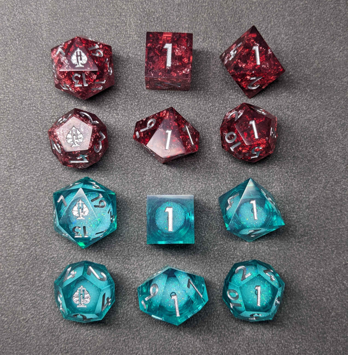 Two sets of dice, each has six shapes. One set is blood red, the other is a silvery transparent blue. 4 of 6 dice in each set lack the Huckleberry logo.