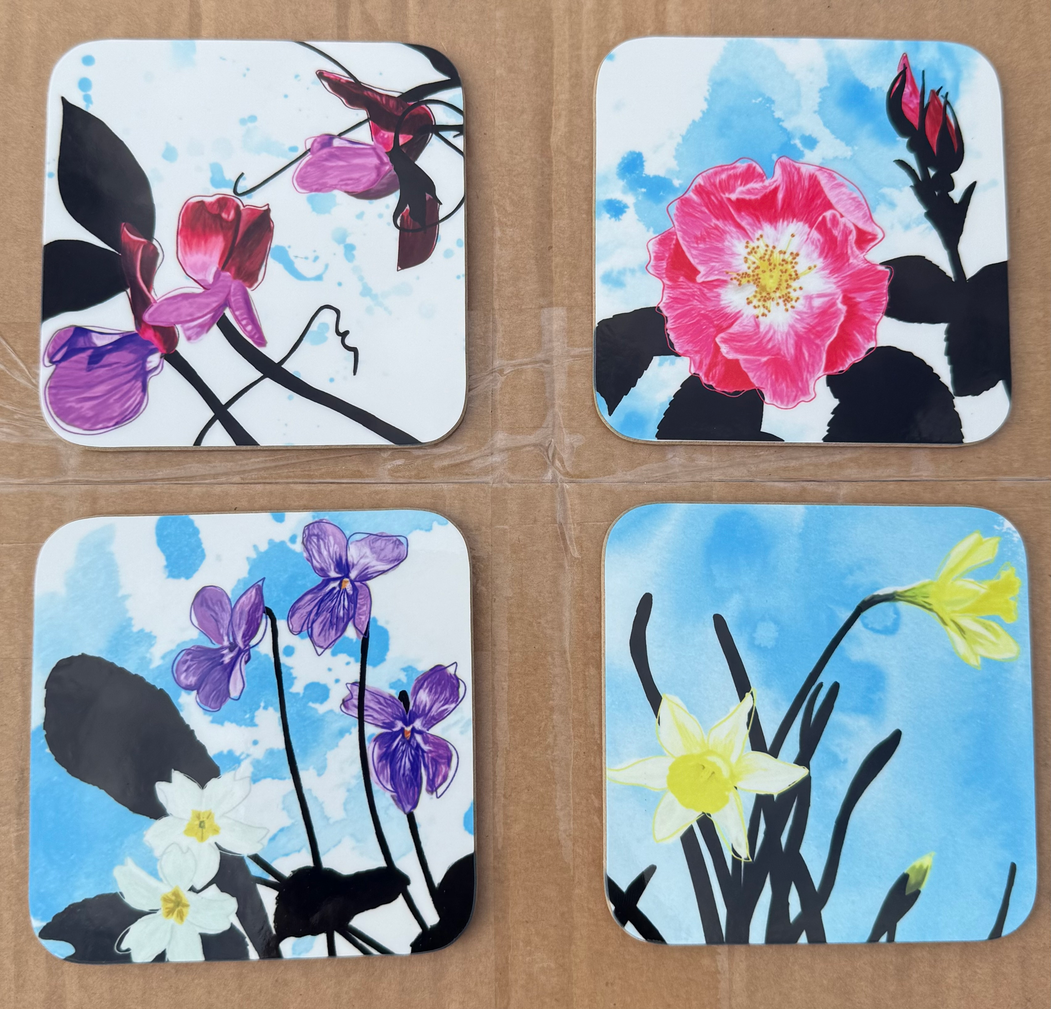 four flower themed coaster samples. Clockwise from top left they show some purple and red sweet peas, a pink wild rose, yellow daffodils and purple violets and white evening primroses.