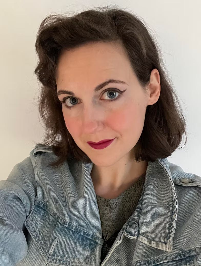  A photo of Georgia, she's a white woman with shoulder-length black curly hair and blue eyes. She's wearing a denim jacket over a jumper and smiling a little 