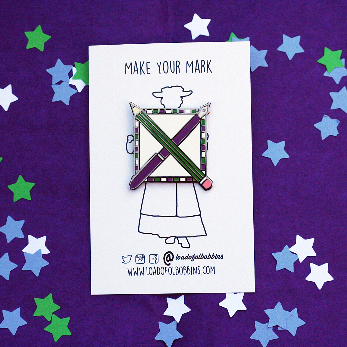 a hard enamel pin with black nickle plating. It is square in shape with a pen and pencil crossing each other to resemble a cross in a voting box. The background is white, the pencil is green and the pen is purple. There's a green, purple and white border around the main box.