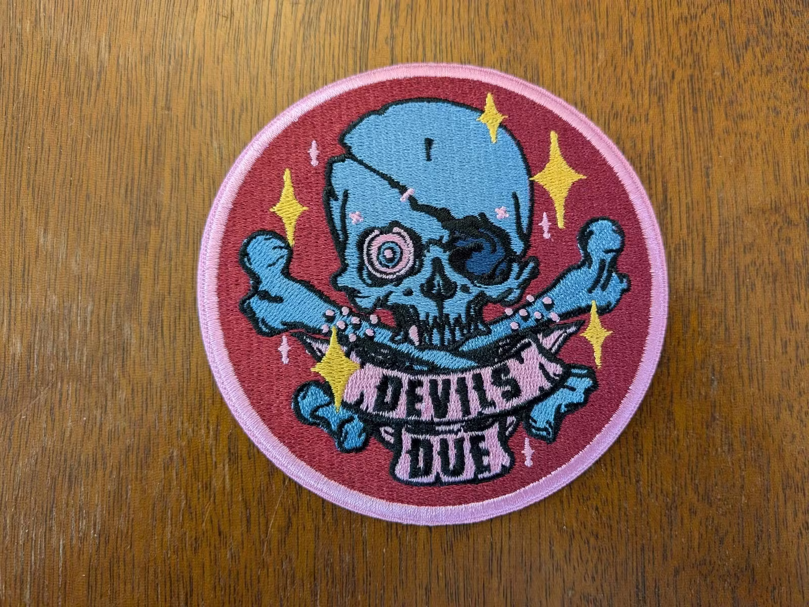  Detail of our Devil's Due patch sample. A blue skull and crossbones over a purple field with yellow and pink stars. A pink banner beneath it reads "Devil's Due." 